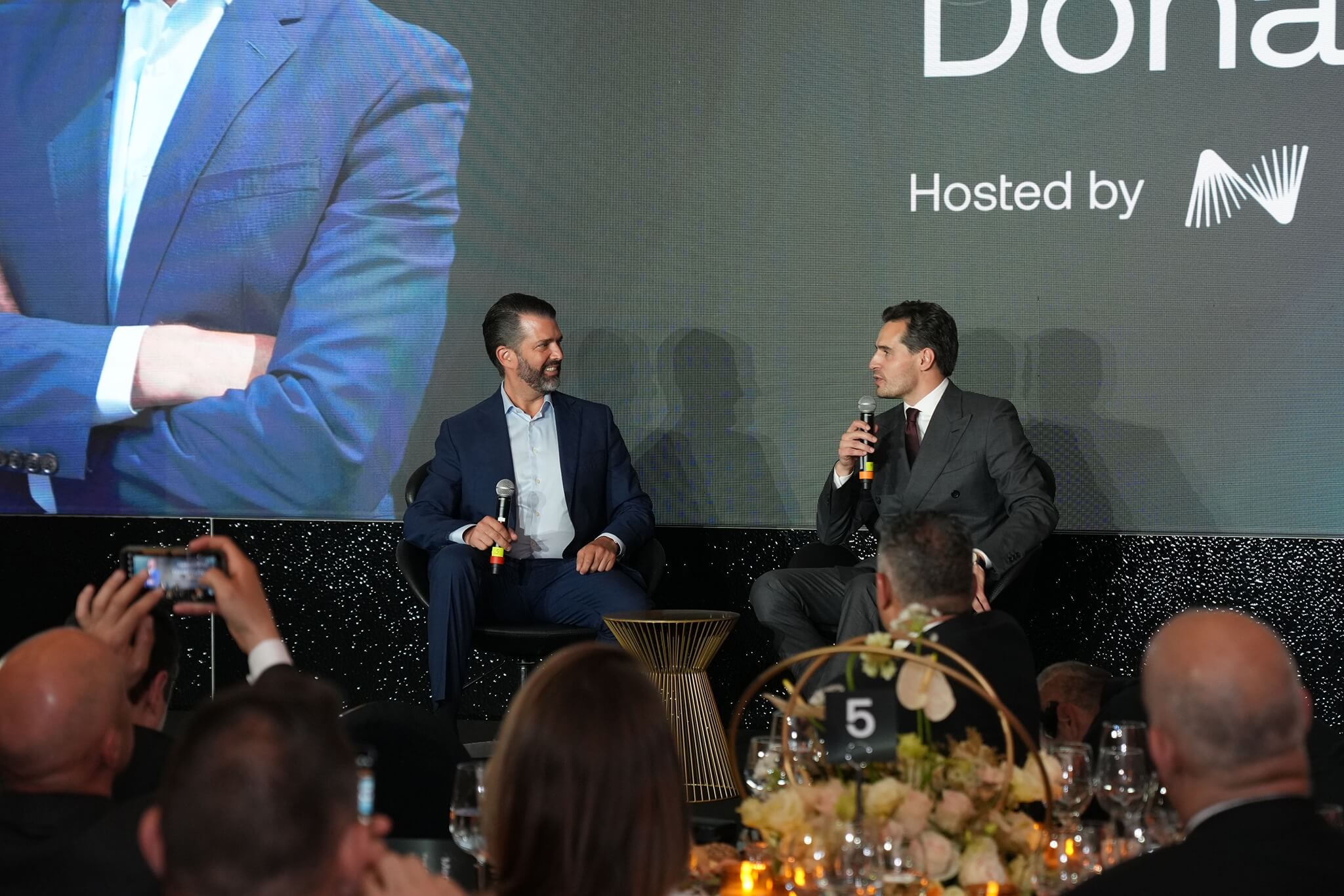 Antoni Trenchev and Donald Trump Jr. sit on stage holding microphones and speaking to each other during a Nexo-hosted event.